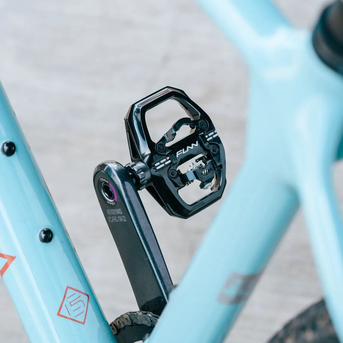 Close-up of black Funn Clast Clip-In Pedals, crafted from die-cast aluminum, attached to the crank arm of a light blue bike with decorative decals visible in the background.