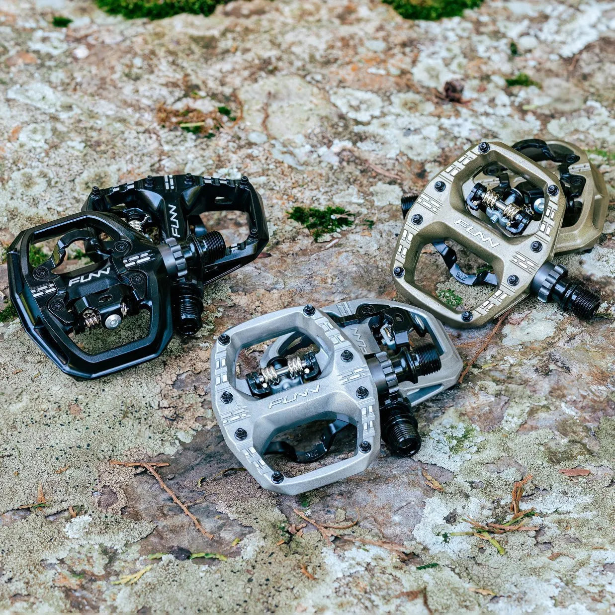 Three pairs of Funn Clast Clip-In Pedals—in black, silver, and bronze—are shown on a rough stone surface with moss and twigs. Each pair features a flat platform design made from durable die-cast aluminum for reliable performance.
