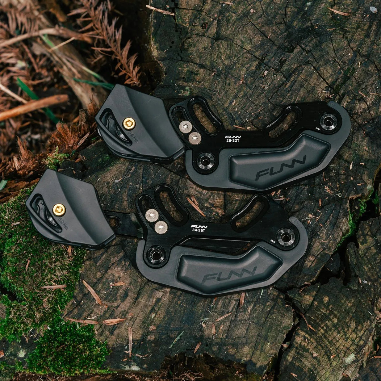 Two black Funn Zippa LG Chain Guides, CNC-machined from alloy, rest on a mossy tree stump surrounded by greenery and leaves in a forest—perfect for high-pivot bikes.