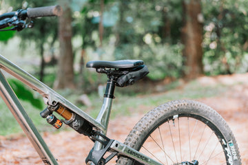 銀色 Commencal 全避震登山車在林道上的側後方視角，黑色 Granite Quiver 工具卷包捲起收在座墊下方