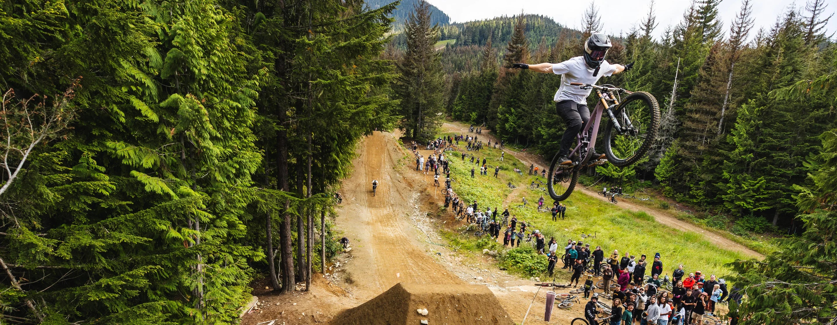 Person on a mountain bike performing a jump in a forested area with spectators watching.
